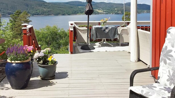 A wooden deck with a view of a lake and mountains, a table with a vase of flowers, and a chair with a floral cushion. - 8 Person Holiday Home in Klokkarstua (Asker)