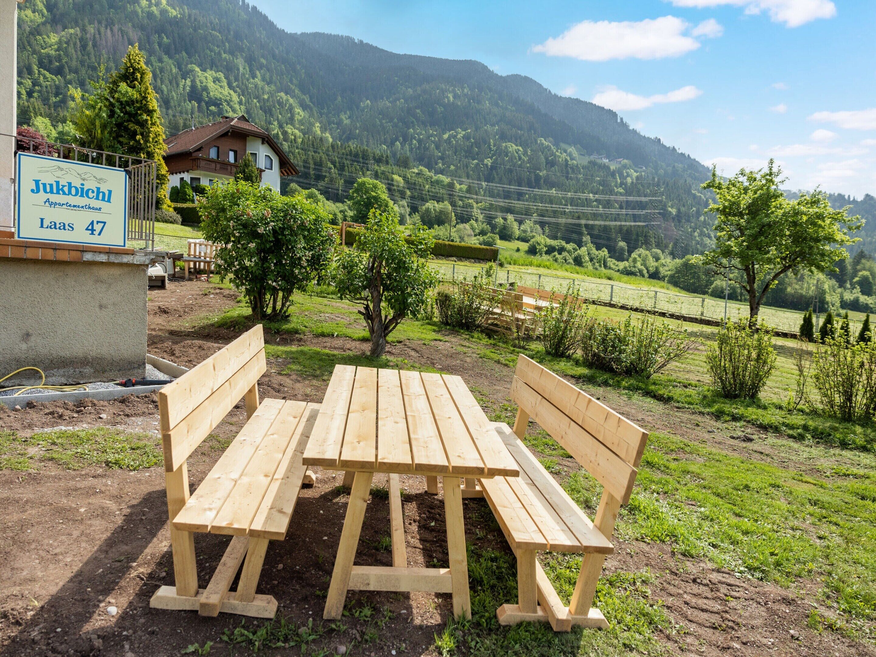 Un banc en bois et une table de pique-nique dans un cadre rural, avec une pancarte indiquant : « Jukbichl Appartementhaus Laas 47 ».