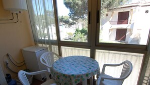 Um quarto pequeno com uma mesa redonda e quatro cadeiras brancas, um aquecedor branco e uma janela com vista para um edifício e árvores.