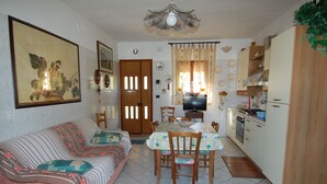 Um espaço de estar compacto com sofá, mesa de jantar, cadeiras, televisão, área de cozinha e uma luminária decorativa no teto.