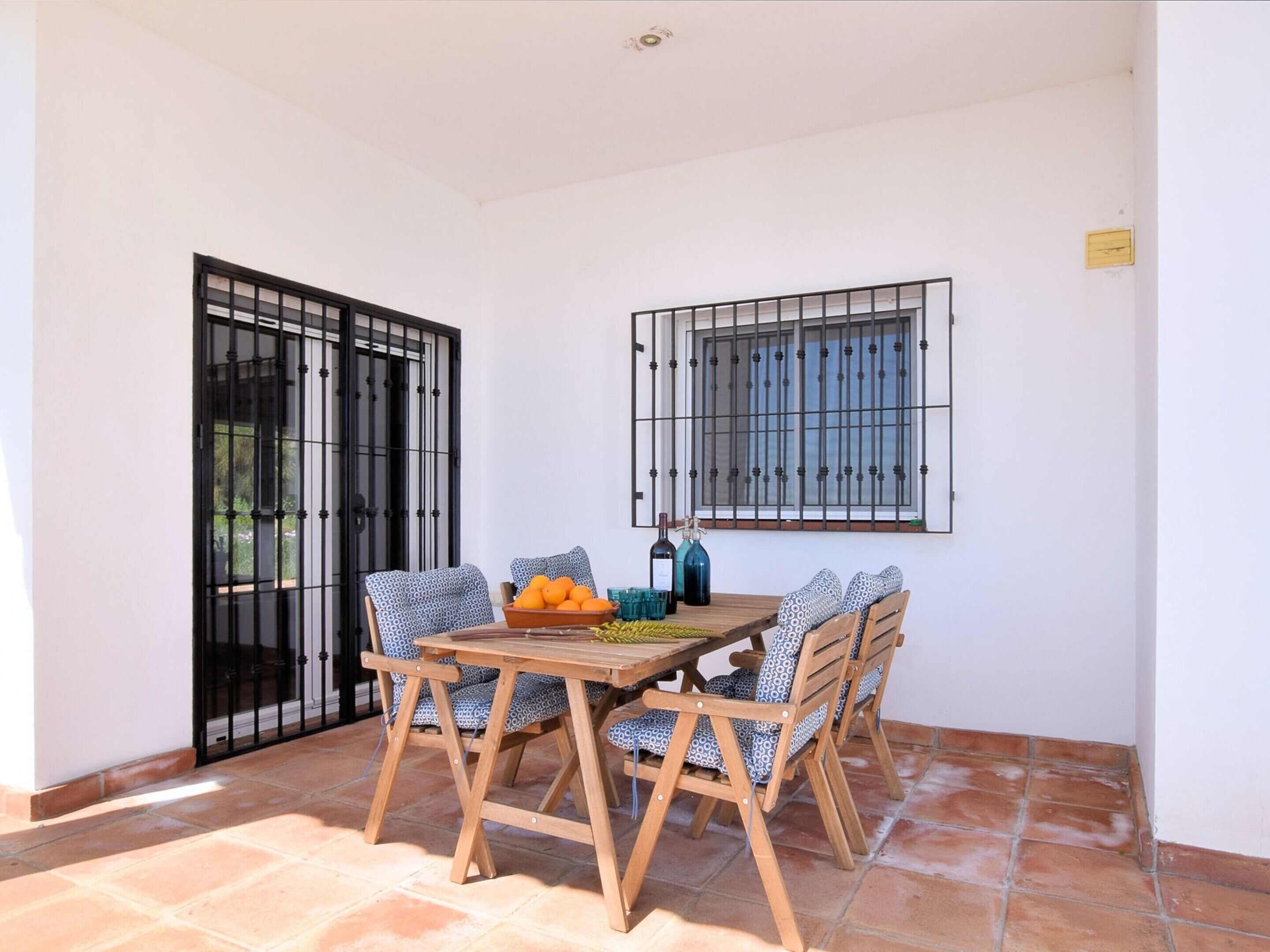 Eine Terrasse mit Holztisch und -stühlen, ein Fenster mit Metallgitter und ein Fliesenboden.