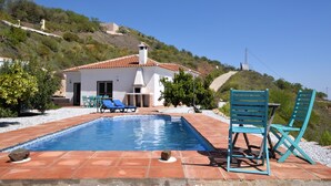 Une piscine entourée de chaises longues bleues, un bâtiment blanc avec un toit recouvert de tuiles rouges, et une colline en arrière-plan.