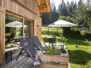A wooden deck with two lounge chairs and a table, surrounded by glass doors and umbrellas.