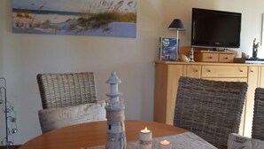 Un salon chaleureux avec une table en bois, des fauteuils en osier, un téléviseur et un tableau représentant une scène de plage.