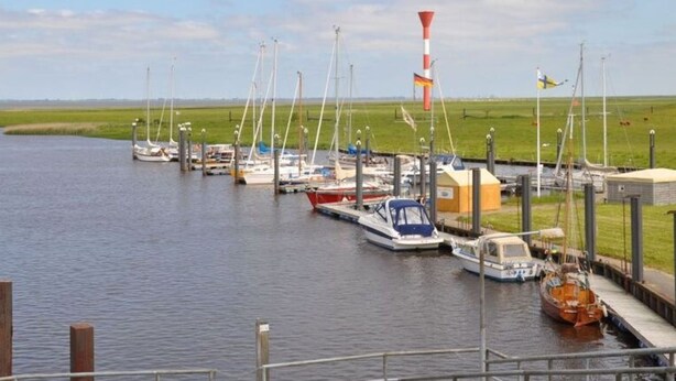 Un port de plaisance où sont amarrés de nombreux bateaux, un mât avec un drapeau, et un paysage verdoyant en arrière-plan.