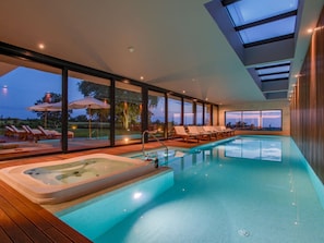 Une piscine intérieure moderne dotée d’un jacuzzi, de chaises longues et de grandes fenêtres offrant une vue sur l’extérieur.