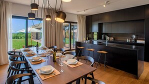 A modern kitchen with dark cabinetry and a central island, a dining area with a wooden table set for six, and large windows offering a view of the outdoors. - Luxury Villa sea View (Porec)