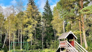 A wooden playhouse with a slide and stairs, surrounded by a grassy area and trees.