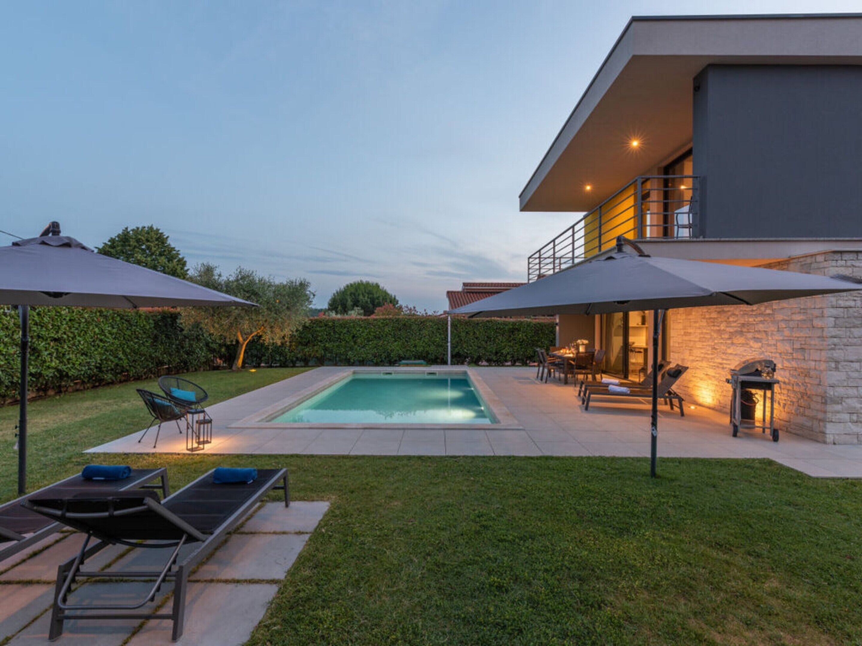 A modern outdoor pool area with lounge chairs and umbrellas, surrounded by a well-maintained lawn and a stone wall.