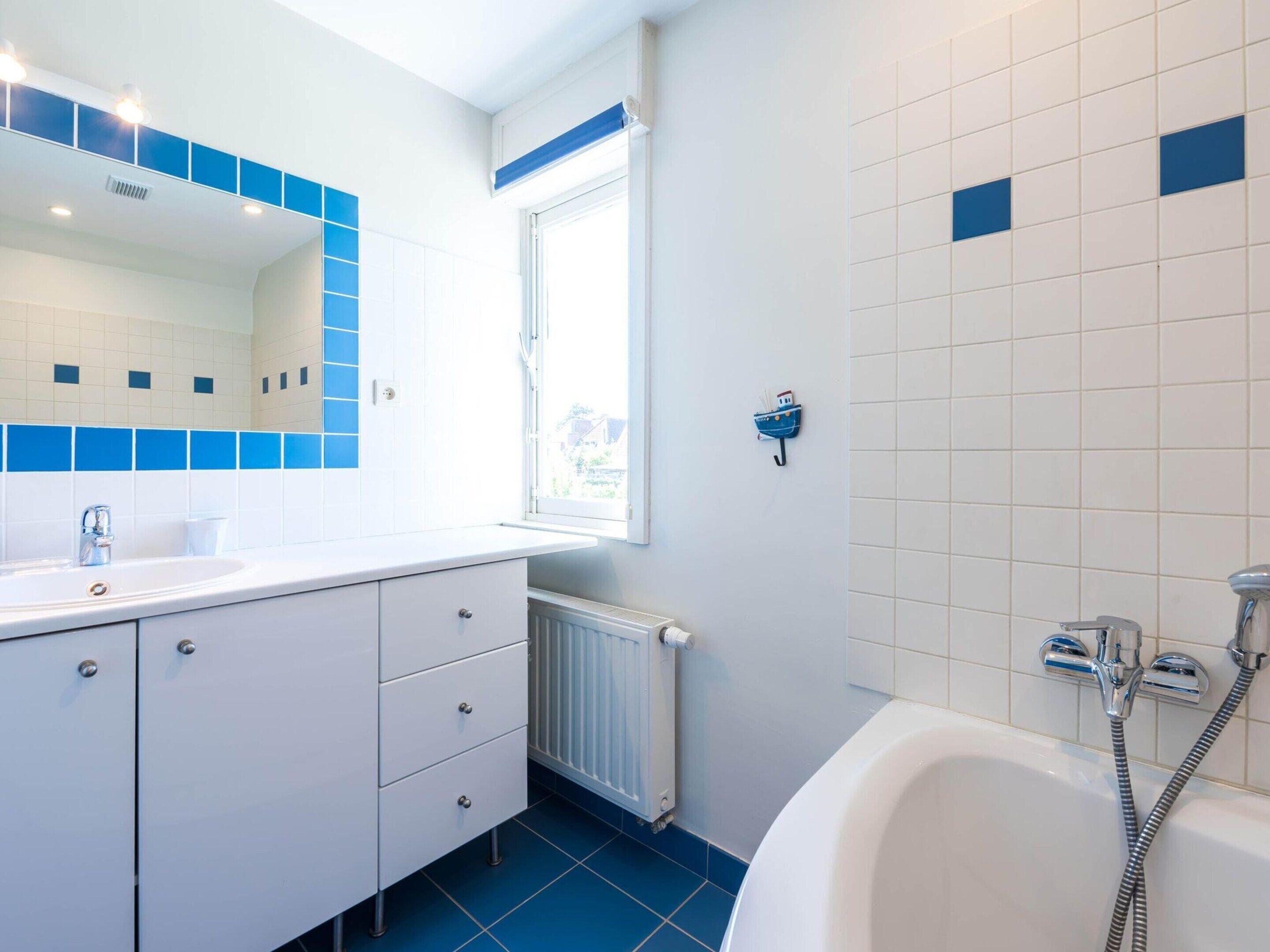 Ein modernes Badezimmer mit einem weißen Waschbecken, einem Spiegel und einer Badewanne. Ein Fenster sorgt für natürliches Licht, und die Wände sind in Weiß und Blau gefliest.