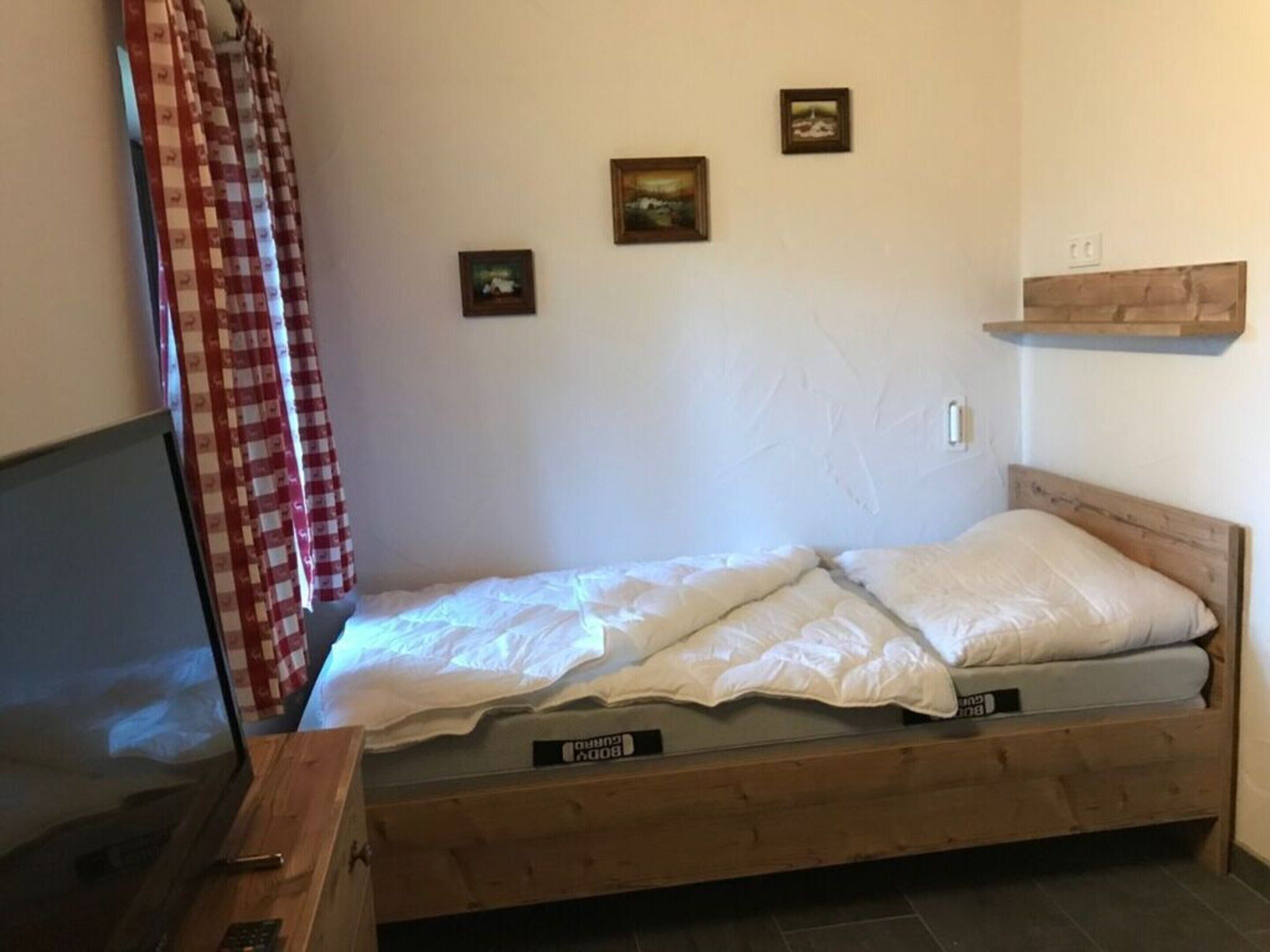 A single bed with a wooden frame and a mattress labeled 'Bedding'. There are three framed pictures on the wall, a red and white checkered curtain, and a wooden shelf above the bed.