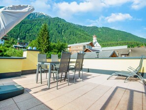 Eine Terrasse mit Sitzgelegenheit im Freien, mit Blick auf einen Berg und ein Gebäude mit rotem Dach.