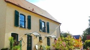 A two-story building with a yellow facade, green shutters, and a tiled roof. There are outdoor seating areas with chairs and tables, and potted plants around the building. - Zonnehof in het Geiseltal (Mücheln (Geiseltal))