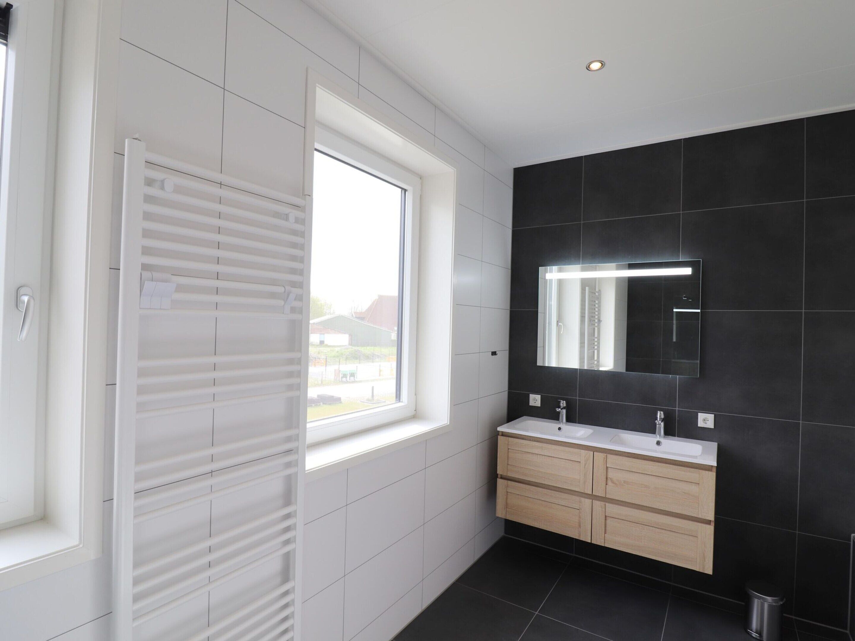 A modern bathroom with a large window, a white vanity with a mirror, and dark tiled walls.