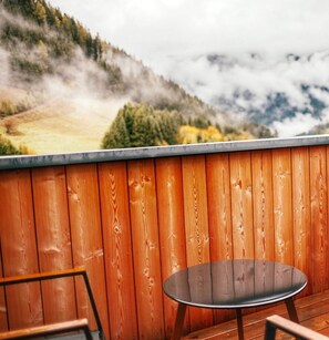 Eine Holzterrasse mit einem Metalltisch und -stühlen, die auf eine gebirgige Landschaft mit Bäumen und bewölktem Himmel blickt.