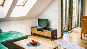 A modern loft apartment with a skylight, a flat-screen TV on a wooden TV stand, a green sofa, a wooden coffee table with a fruit bowl, and a large window with patterned curtains. - Apartment in Hohentauern With Sauna (Hohentauern)