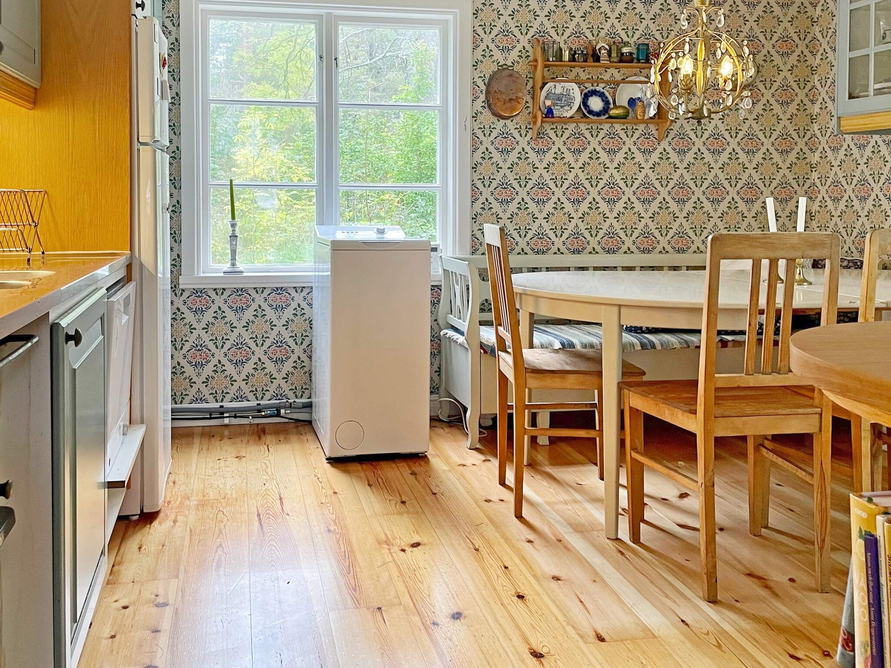 A kitchen with wooden cabinets, a refrigerator, a dining table with chairs, and a window with a view of greenery.