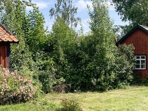 A rustic wooden cabin with a thatched roof, surrounded by dense greenery and flowering bushes.