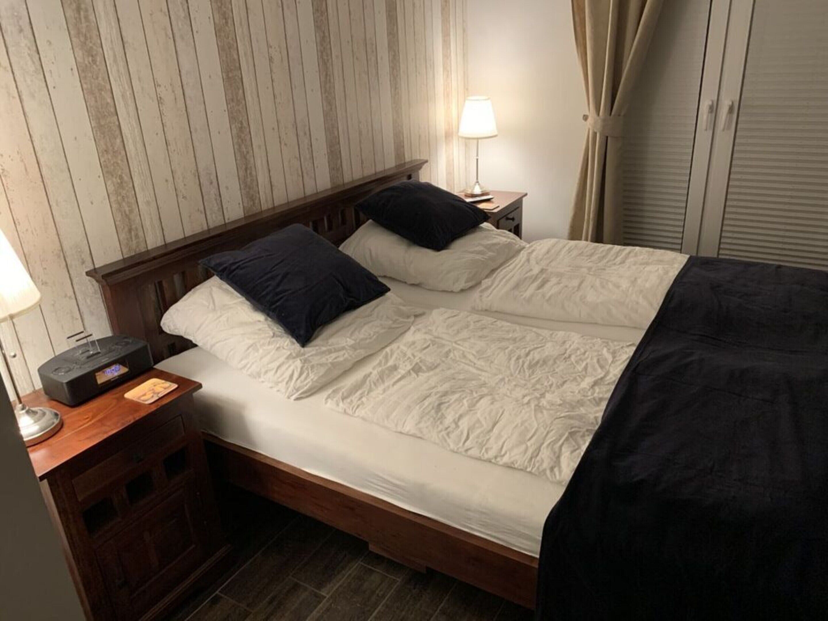 Un lit double avec du linge de lit blanc et des oreillers foncés, une table de chevet avec une lampe et une radio.