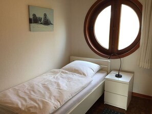 A single bed with a white mattress and a white headboard, a small bedside table with a lamp, a round window with a wooden frame, and a wall-mounted picture of two seals. - Buschsand Lee Comfortable Holiday Residence (Friedrichskoog)