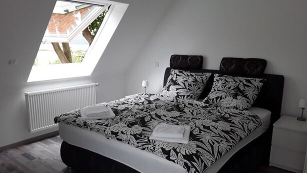 Quarto com claraboia, cama com colcha floral, radiador e mesa de cabeceira com abajur.