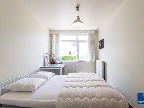 A bedroom with a bed, a bedside table, a window with curtains, a radiator, and a ceiling light. - Oud Klooster 0001 Apartment in De Haan (De Haan)