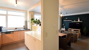 A modern kitchen with a large window, a dining area with a table and chairs, and a dark accent wall in the living room. - Oud Klooster 0001 Apartment in De Haan (De Haan)