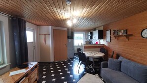 A compact living space with a wooden ceiling, a dining area with a round table and chairs, a sofa, and a kitchen area with cabinets and a clock on the wall. - Kubis Modern Retreat (Wiek)
