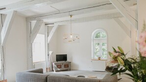 Ett rum i loftstil med synliga takbjälkar, ett stort fönster, en grå hörnsoffa, en tv-apparat placerad på ett träskåp och en hängande lampa.