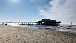 En strand med en brygga som sträcker sig ut i havet, och en byggnad som står på pålar.