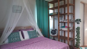 Un dormitorio con una cama bajo una mosquitera, un estante de madera con ropa doblada y una planta en maceta.