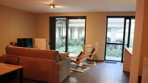 Un soggiorno moderno con un divano, poltrone, una TV e un'ampia porta scorrevole in vetro che dà su un balcone con vista su piante e un edificio.