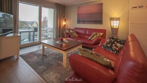 A modern living room with a red leather sofa, a wooden coffee table, and a large window offering a view of the outside. - Holiday Apartment New Love (Zingst)