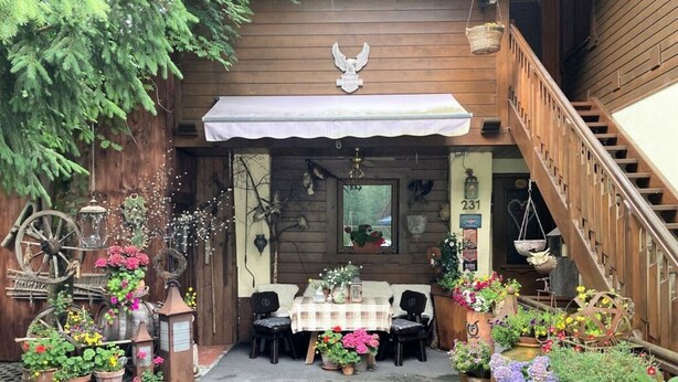 En træbygning med en overdækket terrasse, hvor der er et bord og stole, omgivet af krukker med planter og retroindretning.