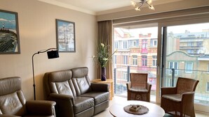 A modern living room with a leather sofa, a round coffee table, and wicker chairs. There are framed pictures on the wall, a floor lamp, and a view of buildings through the large window. - Apartment in Nieuwpoort Near the Beach (Nieuwpoort)