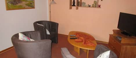 A living room with two armchairs, a wooden coffee table, a television on a wooden stand, a bookshelf with various items, and a painting on the wall.