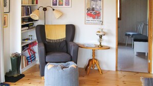 Un saló acollidor amb un sofà armchair gris, un petit reposapiés, una taula auxiliar de fusta amb una làmpada, una prestatgeria i una catifes amb disseny.