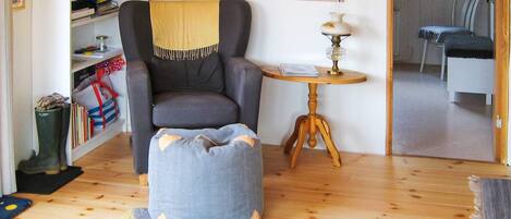 A cozy living room with a grey armchair, a small ottoman, a wooden side table with a lamp, a bookshelf, and a patterned rug.