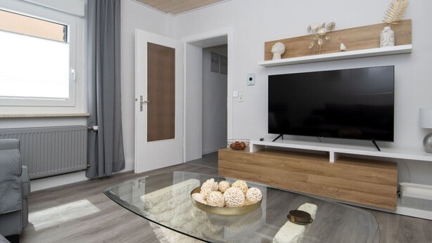 A modern living room with a glass coffee table, a flat-screen TV mounted on a wooden shelf, a grey sofa, and a window with curtains. - Elas Place Modern Retreat (Thür)