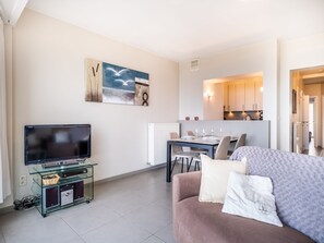 A modern living room with a flat-screen TV, a glass entertainment unit, a sofa with cushions, and a dining area with a table set for four. - Apartment in Belgium Near St-andre Dunes (Koksijde)