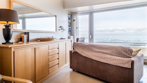 Ein modernes Zimmer mit großem Fenster und Meerblick, einem Holztisch mit Lampe sowie einem gemütlichen braunen Sofa.