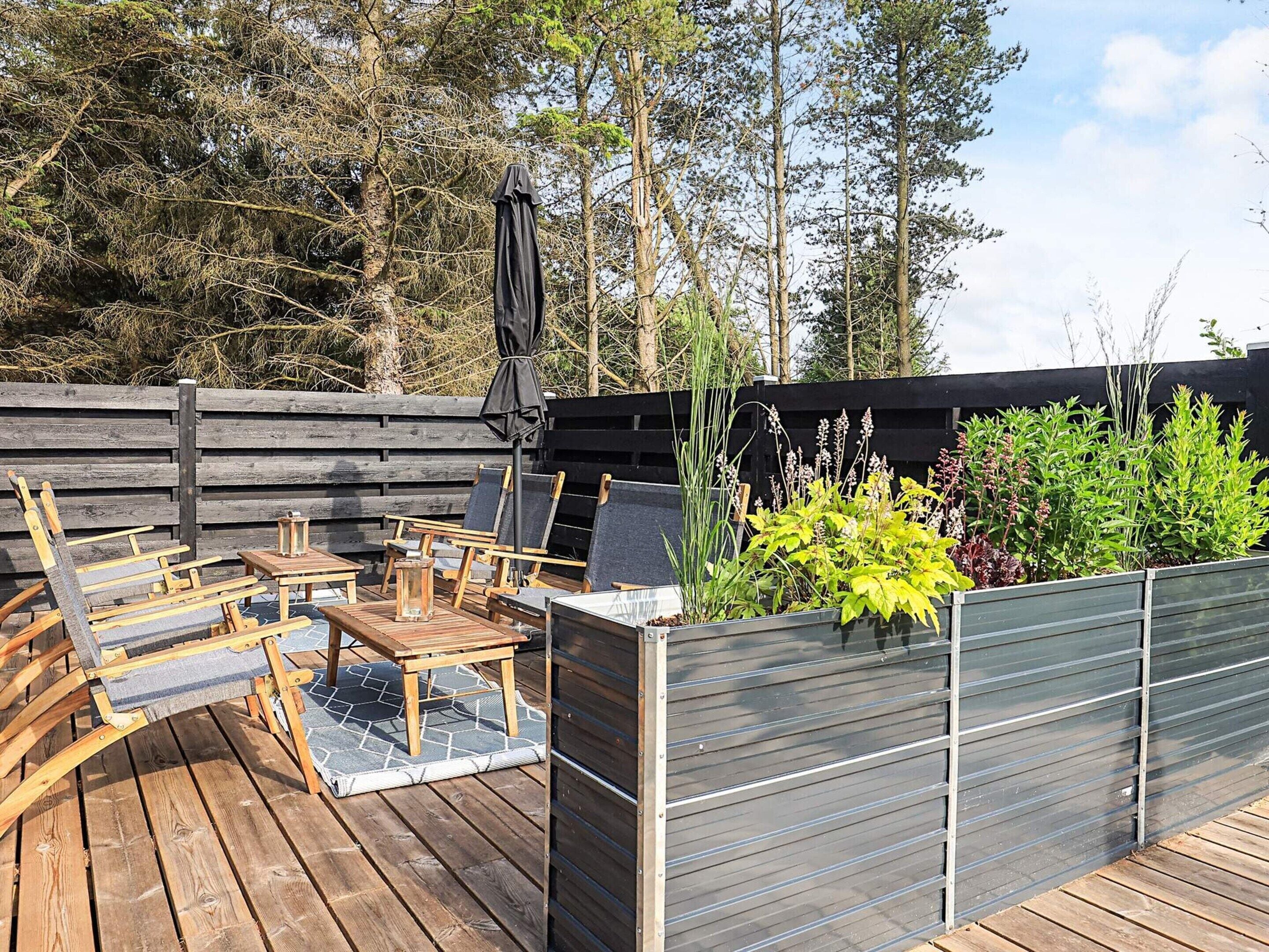 Una terraza de madera con mobiliario exterior, una parrilla y una jardinera con diversas plantas.