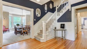 A spacious room with a wooden floor, a dining area with a table and chairs, a staircase with white railings, and a door with a welcome sign. - The Avenue! Historic Mansion in DT Manitou Springs (Manitou Springs)