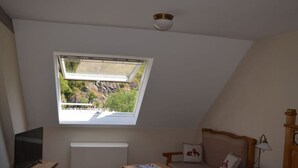 Une chambre avec un plafond en pente, une fenêtre donnant sur la verdure, un lit recouvert d’une courtepointe à carreaux, une table de chevet en bois et une chaise agrémentée d’un coussin à motif de renard.