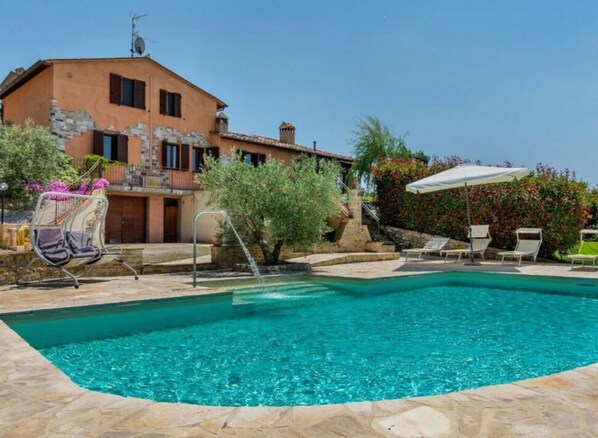A swimming pool with a stone border, a lounge area with chairs, and a building with a stone facade.