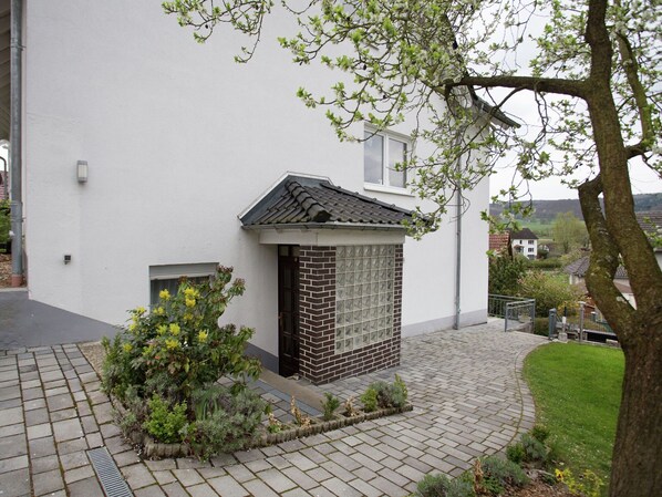 A white building with a small brick structure, a paved walkway, and a tree with yellow flowers. - Schone Ferienwohnung mit Terrasse in Homberg (Homberg)