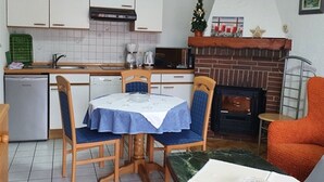 A compact kitchen with white cabinets, a dishwasher, and a microwave. There's a dining table with blue chairs and a small orange armchair. A fireplace with a brick surround is visible, and a small Christmas tree is on the mantel.