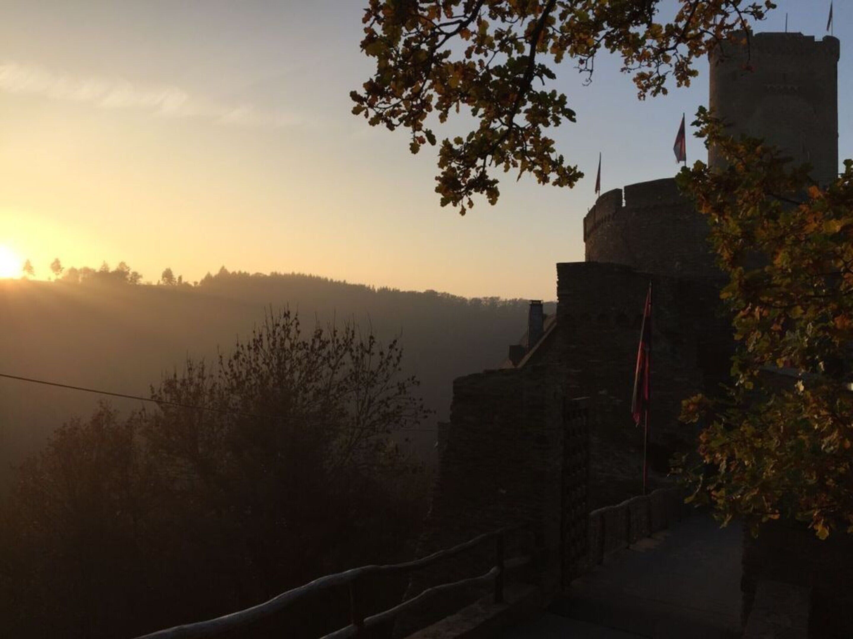 A stone building with a flag, surrounded by trees and a hilly landscape during sunset.