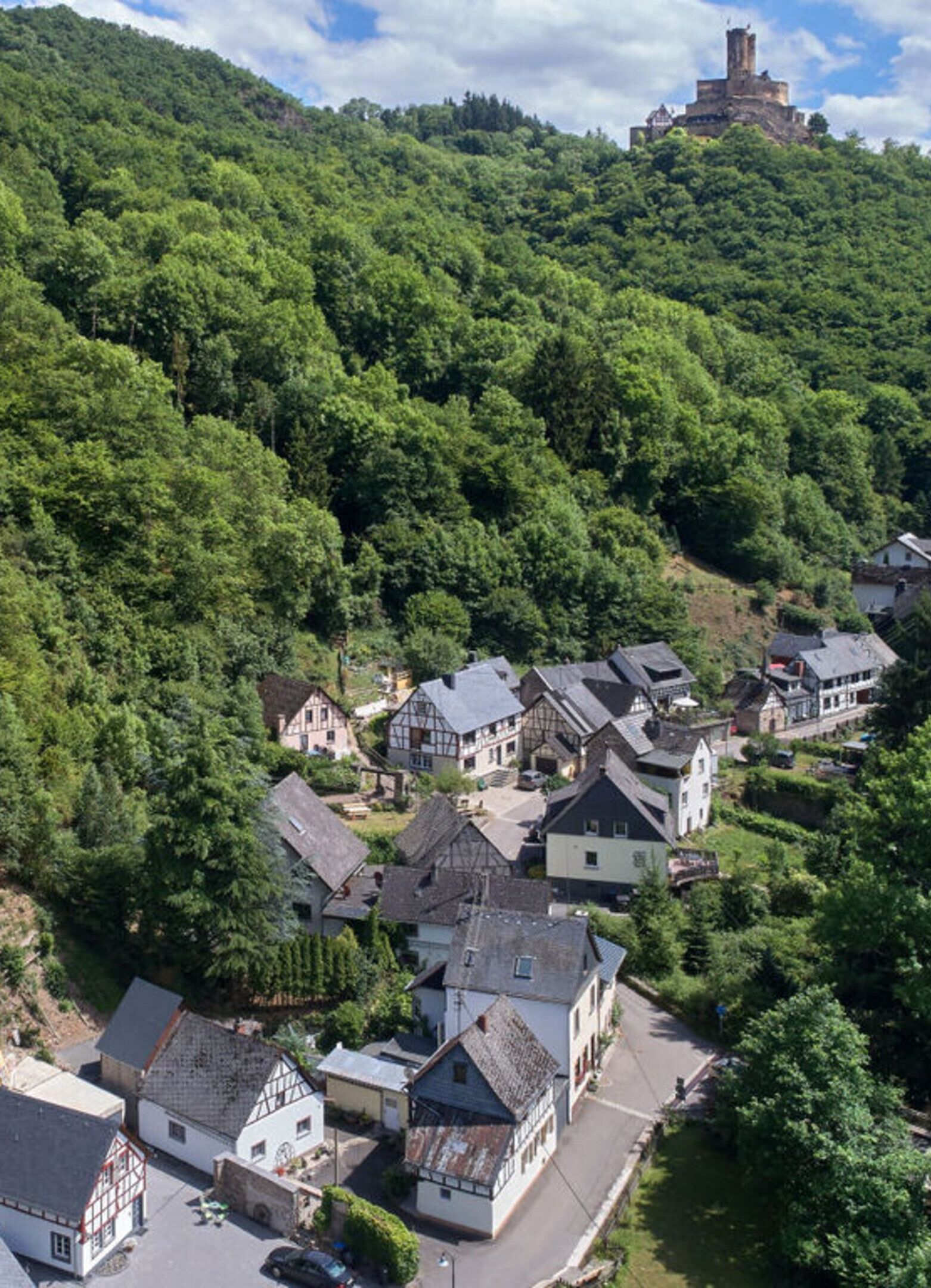 Uma aldeia com casas tradicionais de enxaimel aninhada numa encosta arborizada, um castelo imponente no topo e uma estrada clara que serpenteia pela aldeia.