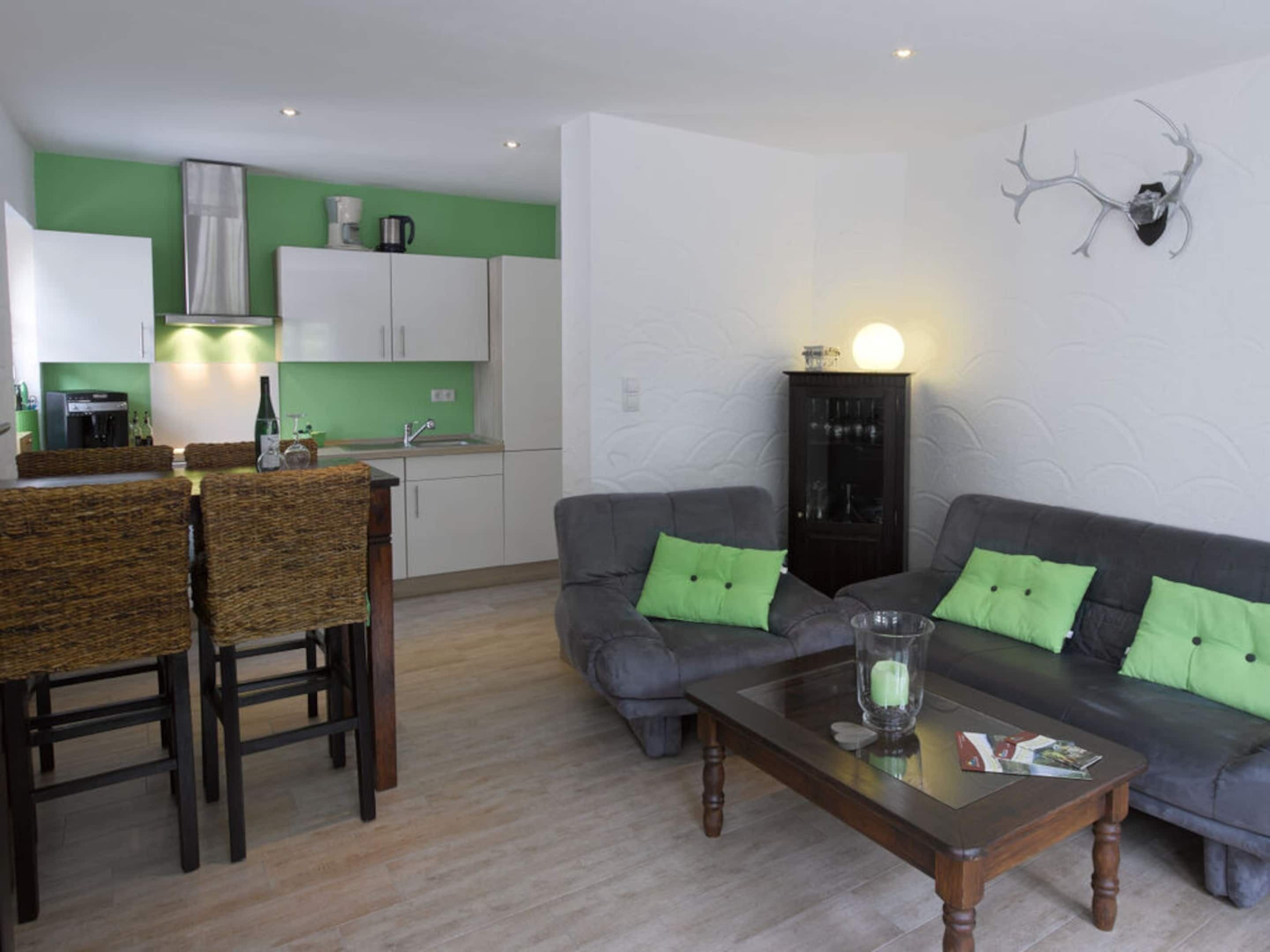 Ein kompakter Wohnraum mit Küchenzeile, einem Essbereich mit Korbstühlen, einem grauen Sofa mit grünen Kissen und einem Glastisch.